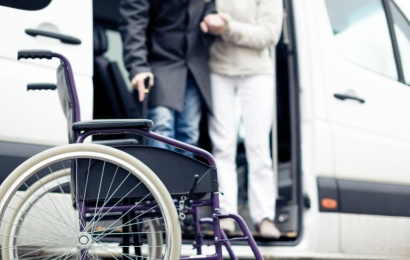 Neįgaliojo vežimėlis prie balto mikroautobuso ir įlipančiam žmogui padedantis asmuo