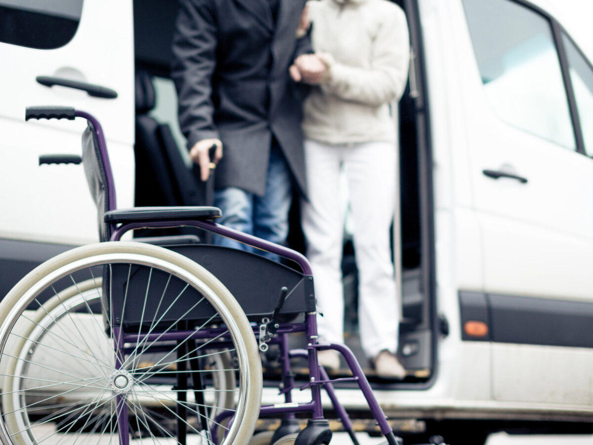 Neįgaliojo vežimėlis prie balto mikroautobuso ir įlipančiam žmogui padedantis asmuo
