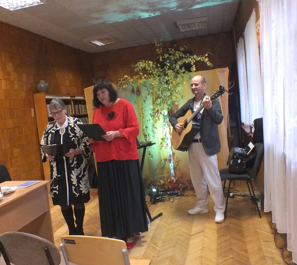 Dvi moterys skaito iš aplankų, šalia vyras groja gitara Vankiškių bibliotekoje.